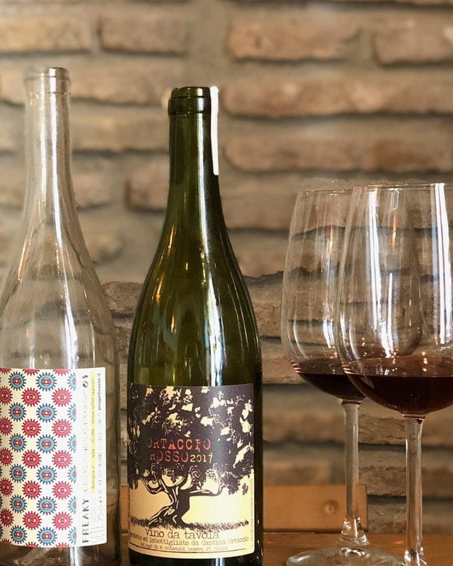 Two wine bottles, one with a colorful circular pattern and one labeled Ortaccio Rosso 2017, next to two glasses of red wine.