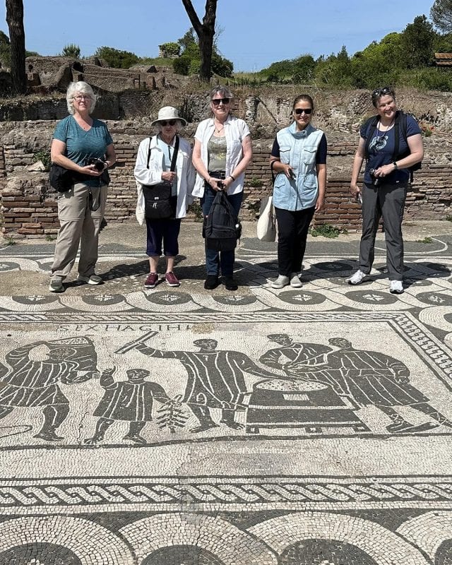 Five women stand in a row behind a large black-and-white ancient mosaic depicting figures at a Roman port.