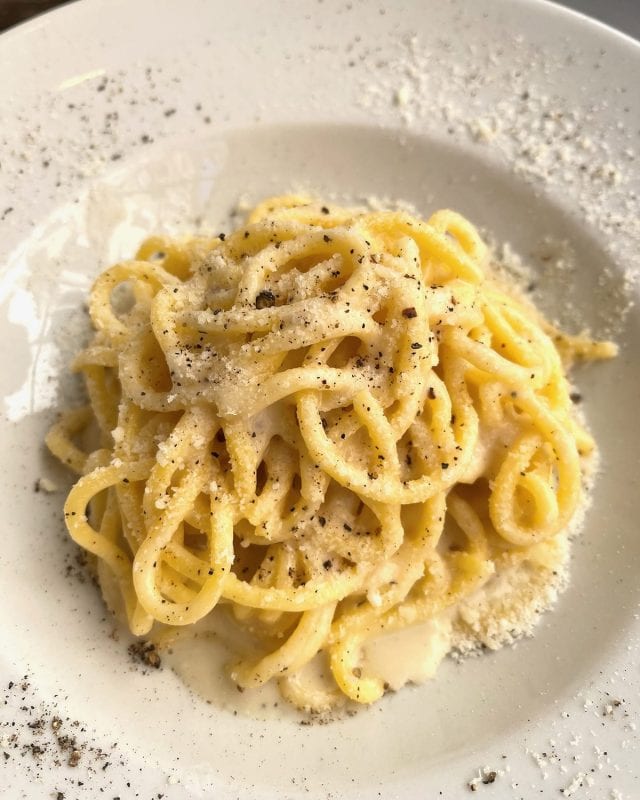 A mound of spaghetti coated in a creamy sauce, topped with grated cheese and cracked black pepper in a shallow bowl.