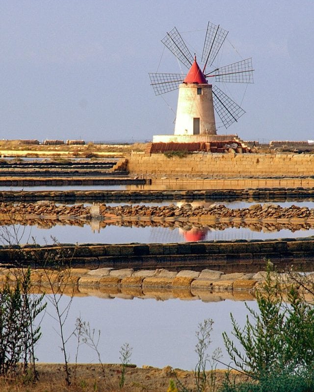 A windmill with a red roof at a salt works, reflected in the surrounding salt pans.