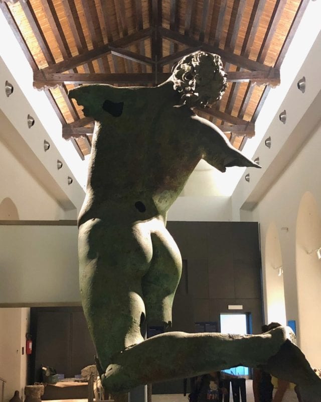 Back view of a bronze statue of a kneeling male figure with missing arms, displayed in a museum.