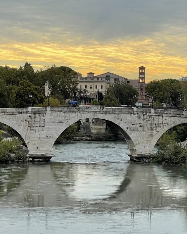 A stone arch bridge crossing a river in Rome at sunset, with a church bell tower in the background.