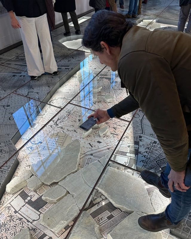 A man in a brown jacket uses his smartphone to photograph a glass-covered floor map with stone fragments placed on top.