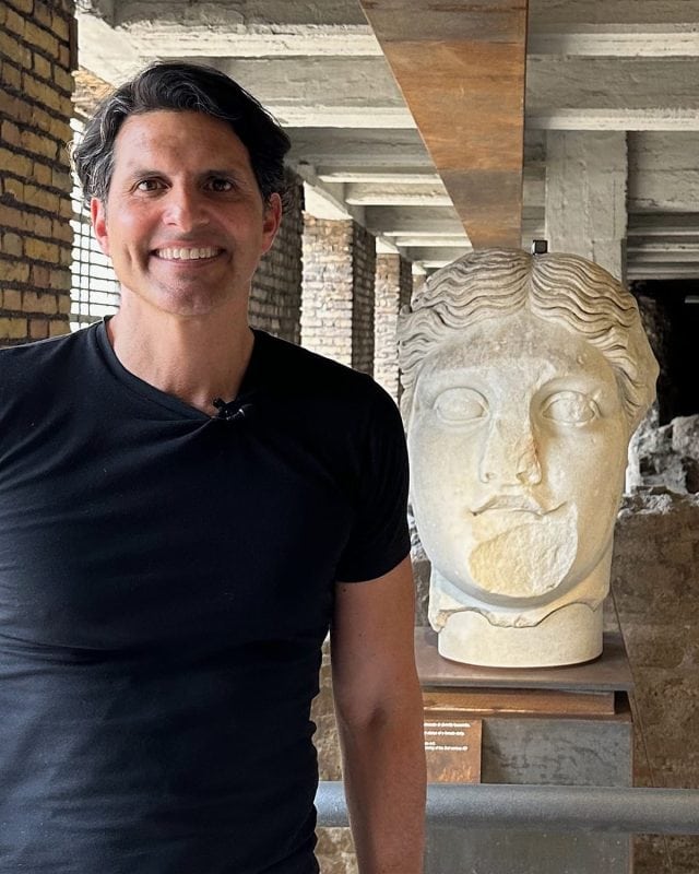 A man in a black t-shirt standing next to a large stone sculpture of a human head in a colonnade.