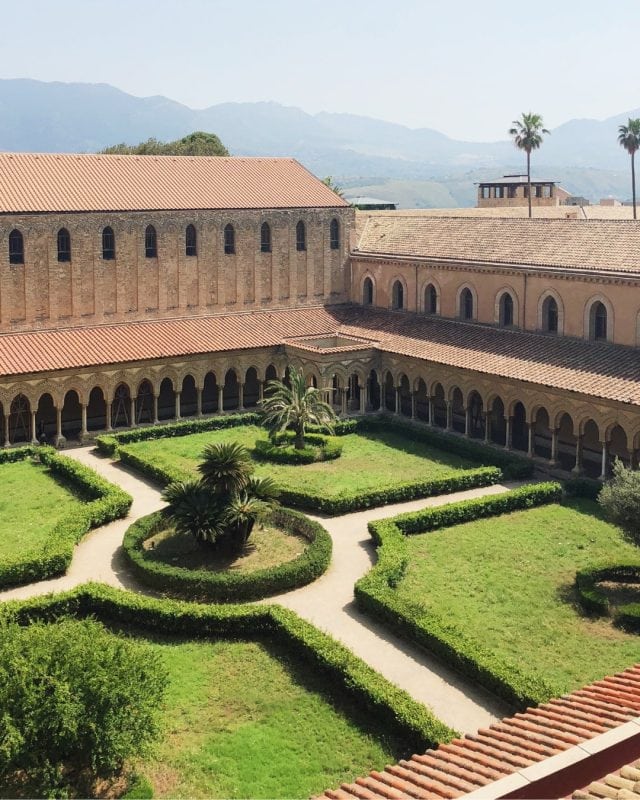 A cloister garden with manicured hedges and palm trees, surrounded by an arched stone arcade and terracotta-roofed buildings.