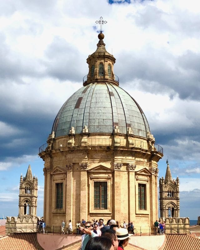 The dome of Palermo Cathedral topped with a cross, with smaller towers in the background and a group of tourists on the terrace below.