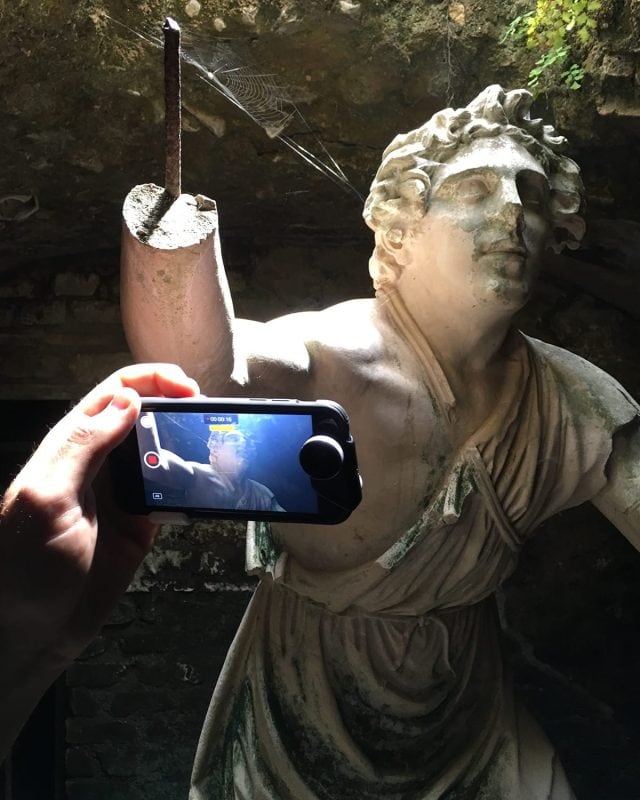 A person holding a smartphone captures a photo of a weathered stone statue of a man with a missing arm.