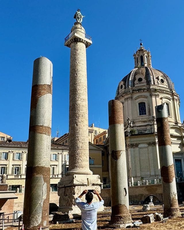 A person photographs Trajan’s Column at the Roman Forum, with the Church of the Most Holy Name of Mary in the background.