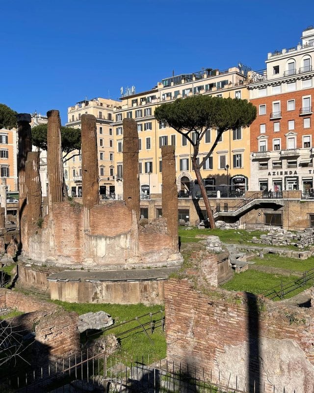 Ancient brick ruins with tall columns at Largo di Torre Argentina in Rome, surrounded by modern city buildings.