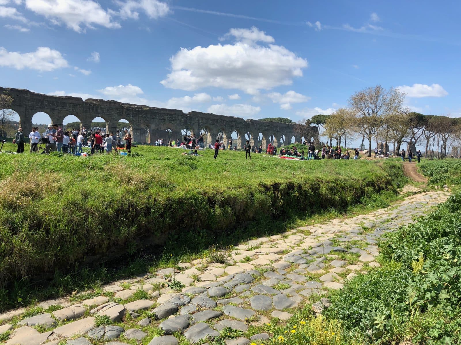 A group of people gather on grass near ancient stone arches and a cobblestone path under a blue sky with scattered clouds.