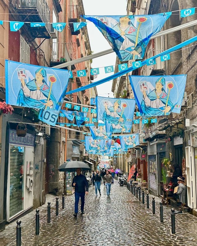 Narrow cobblestone street decorated with blue flags featuring a saint, with pedestrians walking beneath them.