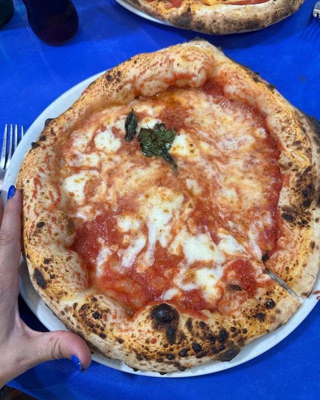 A margherita pizza with melted mozzarella and tomato sauce on a white plate atop a blue tablecloth.