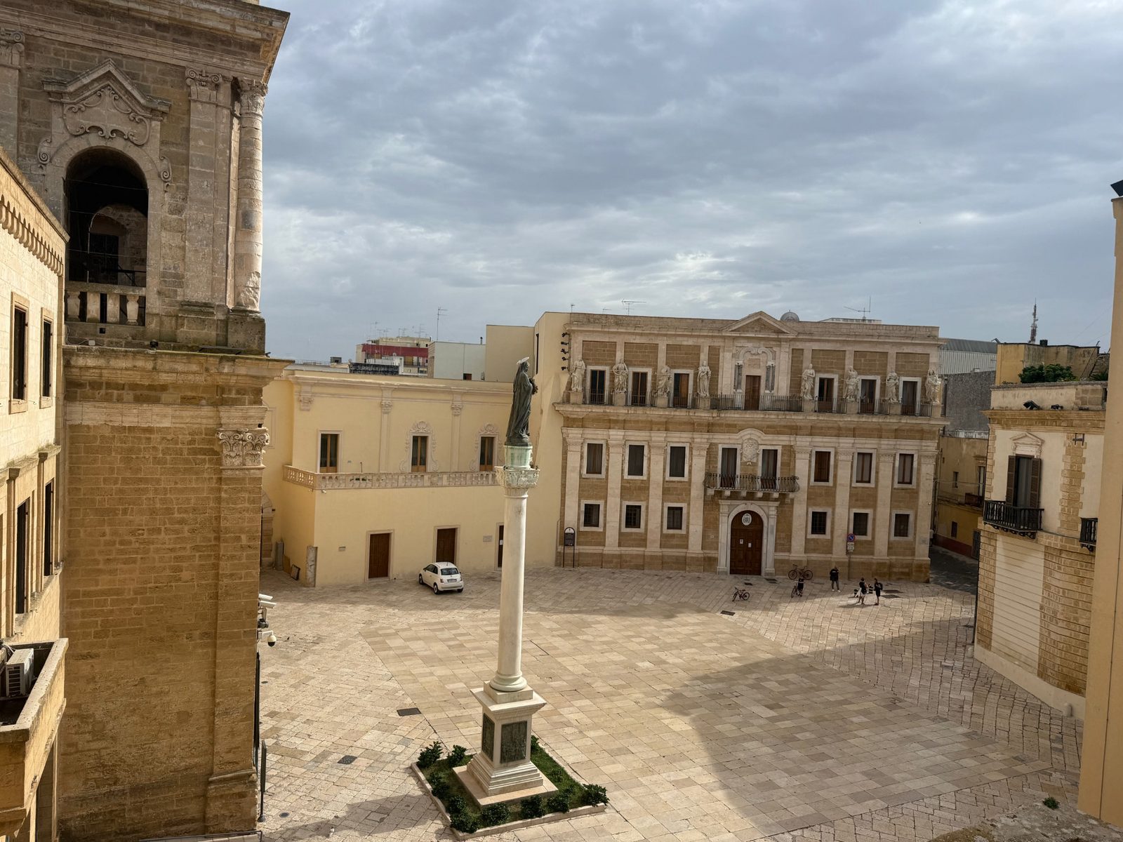 A stone plaza in Lecce, Italy, featuring a central column with a statue, surrounded by historic buildings under a cloudy sky.