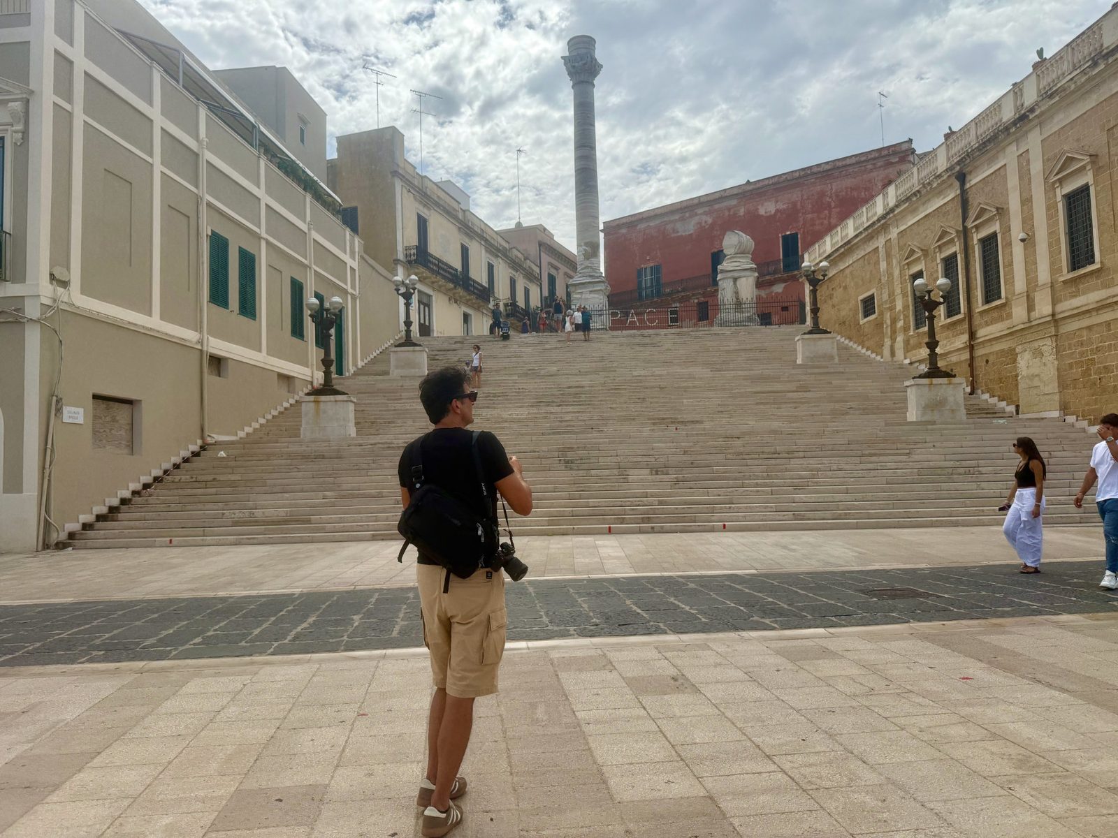A tourist with a camera stands at the base of the wide stone staircase leading up to the Colonna della Libertà in Ostuni, Italy.