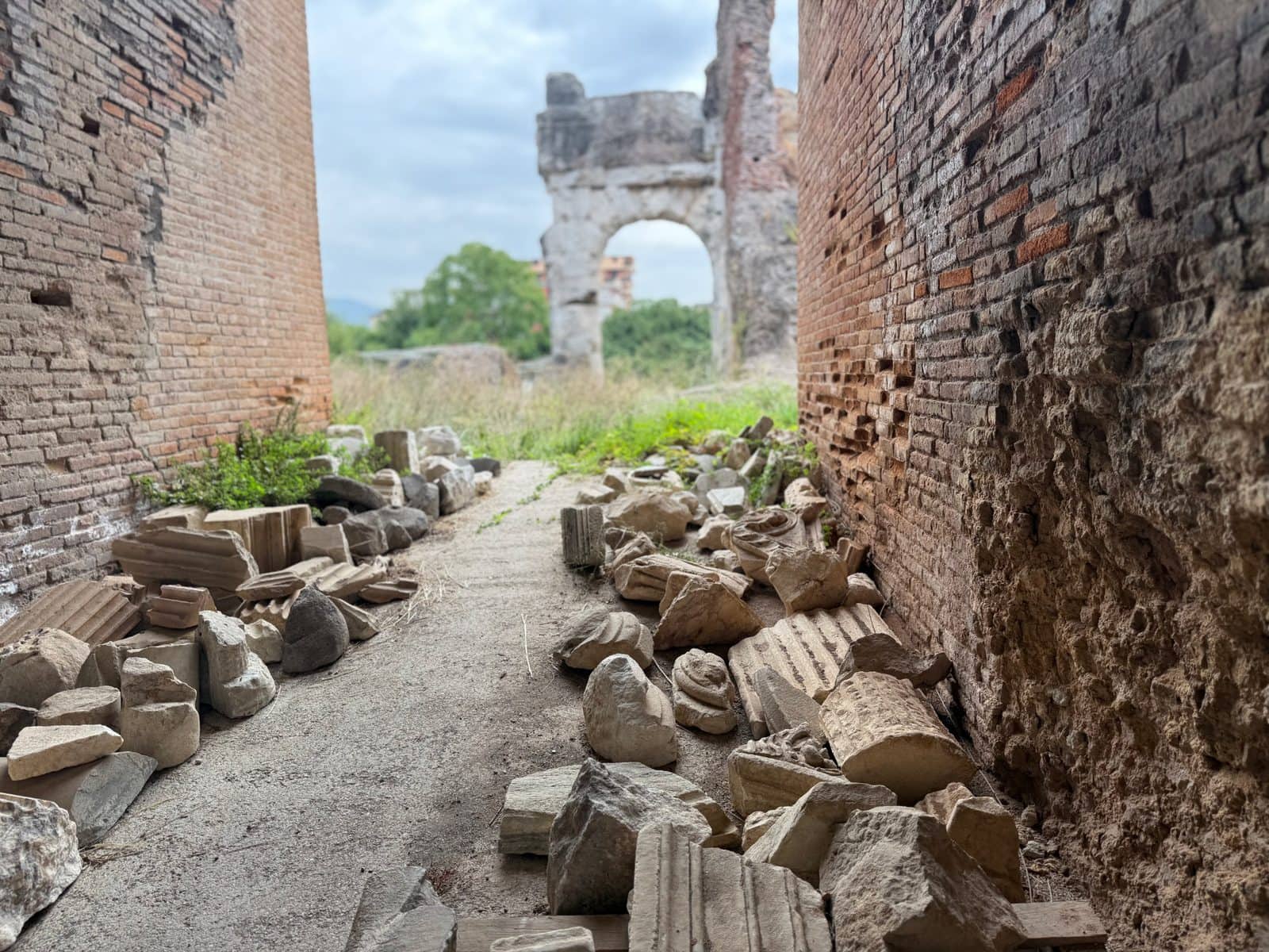 A dirt path between two brick walls leads toward ancient stone ruins and a distant archway.