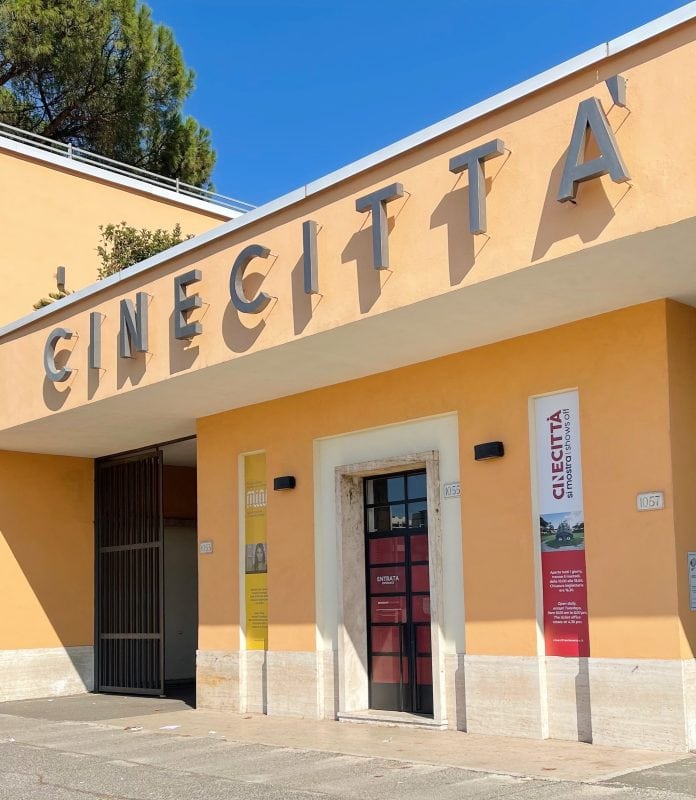 Entrance of Cinecittà studio in Rome with large sign above the door, posters on the wall, and a gated entryway.