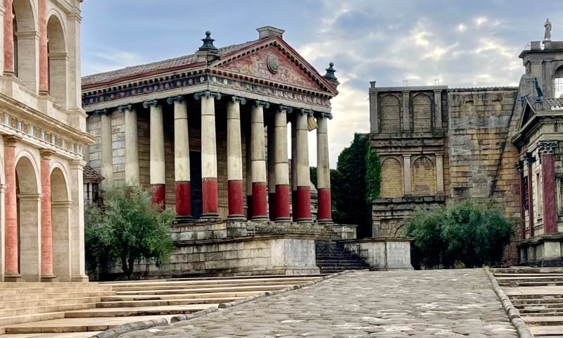 A stone path leads to ancient Roman-style buildings with columns, red and beige walls, and arched windows under a cloudy sky.