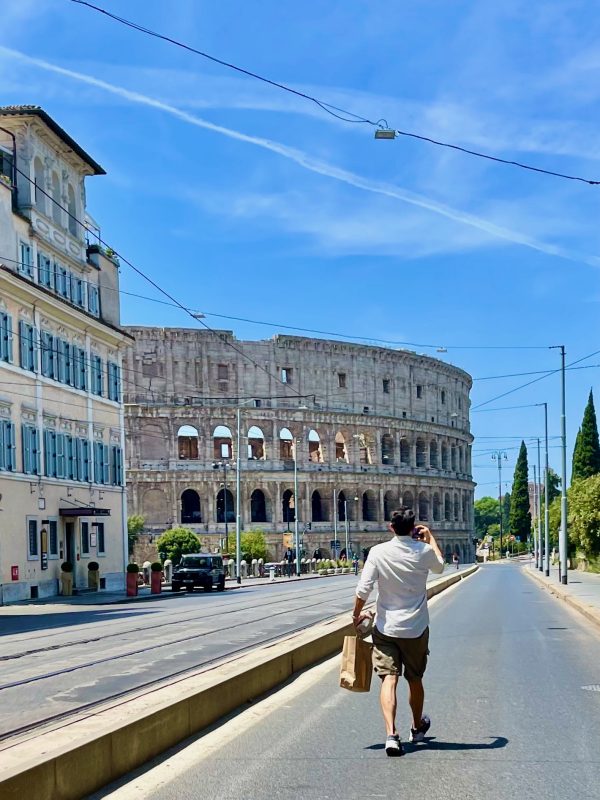 A man walks along a sunny street near the ancient Roman amphitheater in Nîmes, France, holding a paper bag and touching his head.
