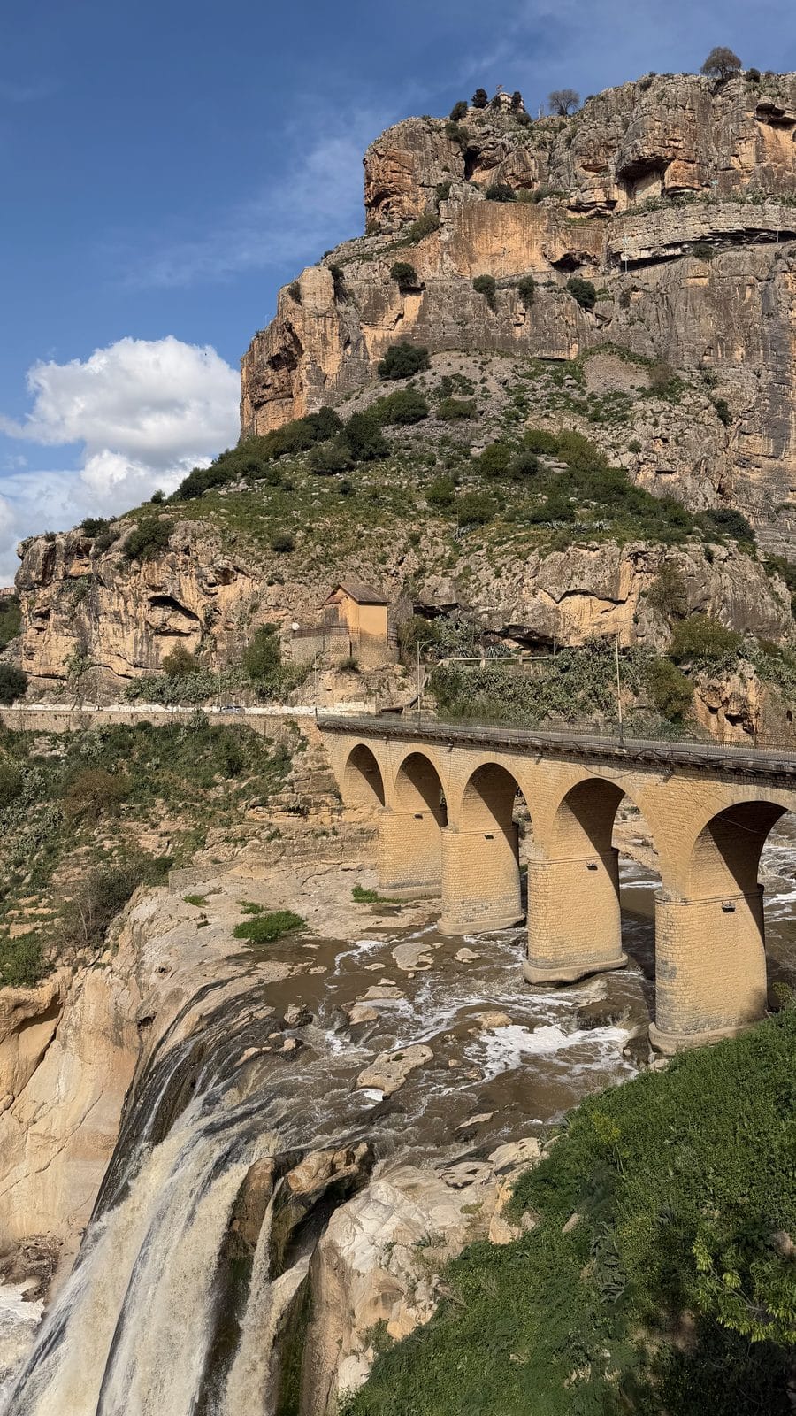 A stone arch bridge crossing a river above a waterfall, set against a steep, rocky cliff with a small building on the hillside.