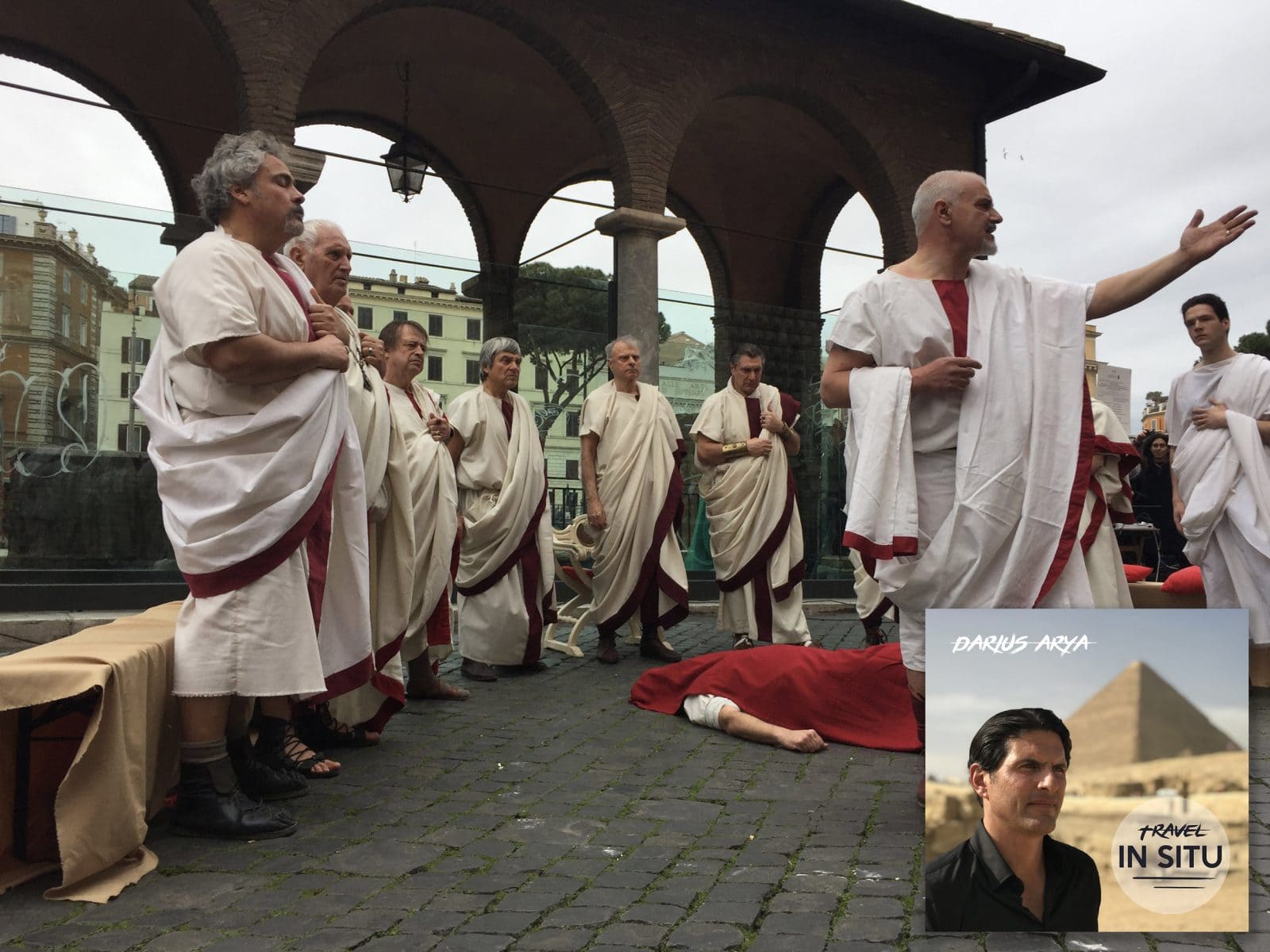 A group of people in Roman togas perform outdoors; one person lies on the ground. An inset shows a man by a pyramid with text “Darius Arya: Travel In Situ.”.