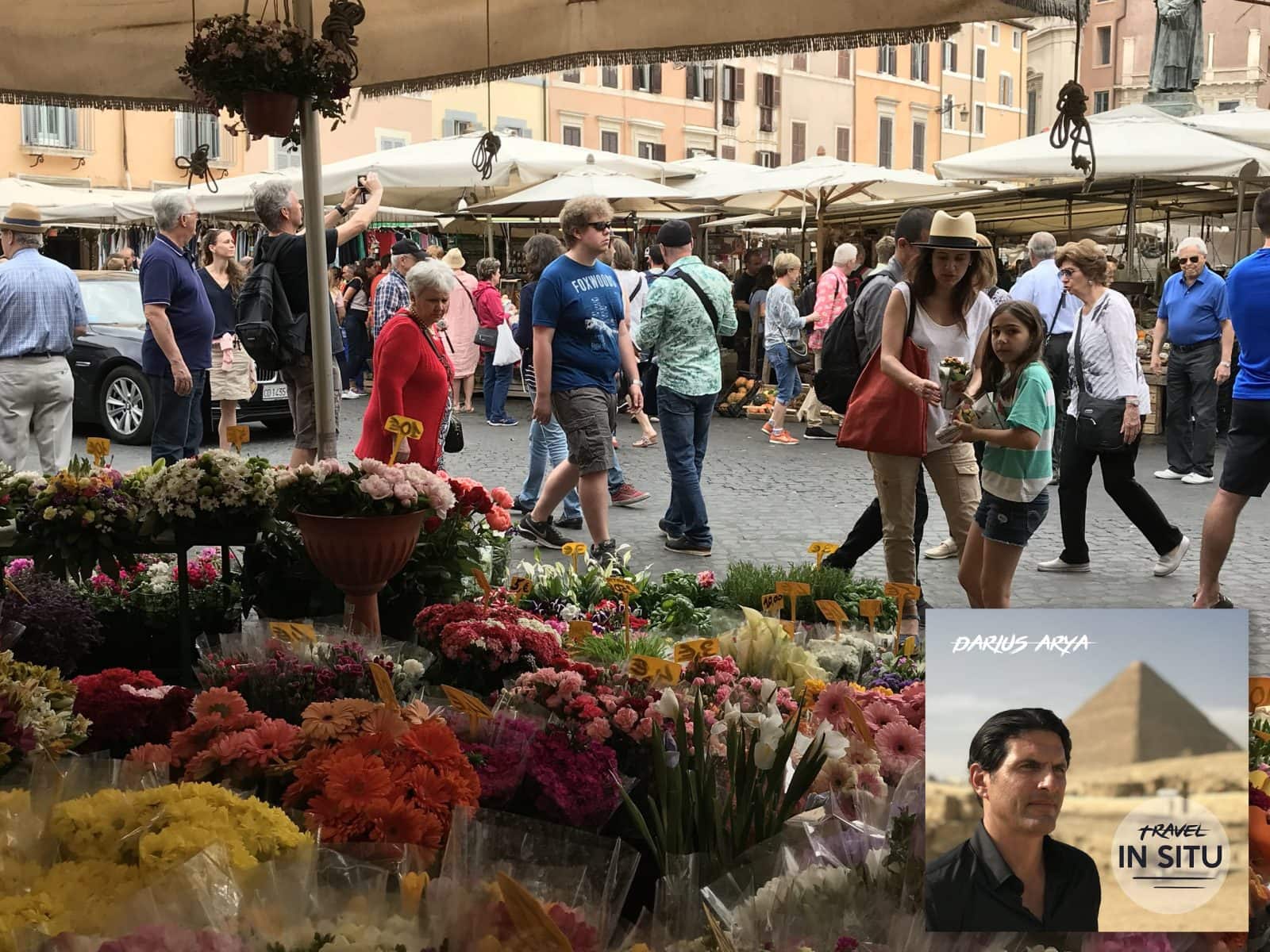 A busy outdoor flower market with colorful blooms in the foreground and people walking among market stalls in a European square; an inset of a man and the text “Travel in Situ” appears.