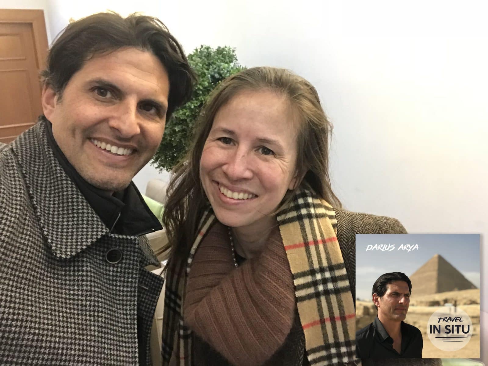 Two people smiling indoors; a poster in the corner shows a man with a pyramid in the background and the text “Darius Arya Travel In Situ.”.