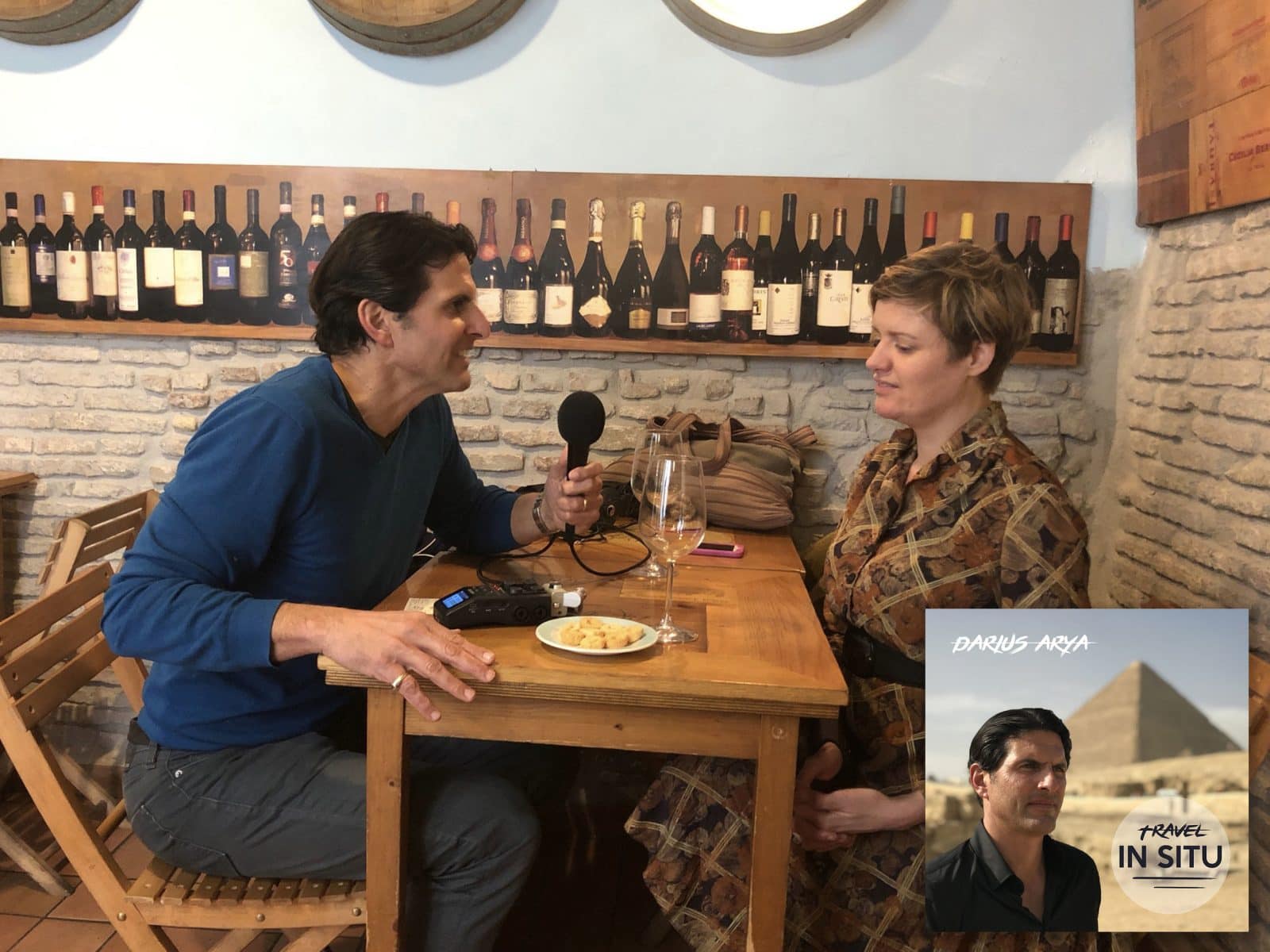 Two people sit at a small table in a wine bar, one holding a microphone in an interview. Travel podcast graphic with text “Darius Arya Travel In Situ” appears in the lower right corner.