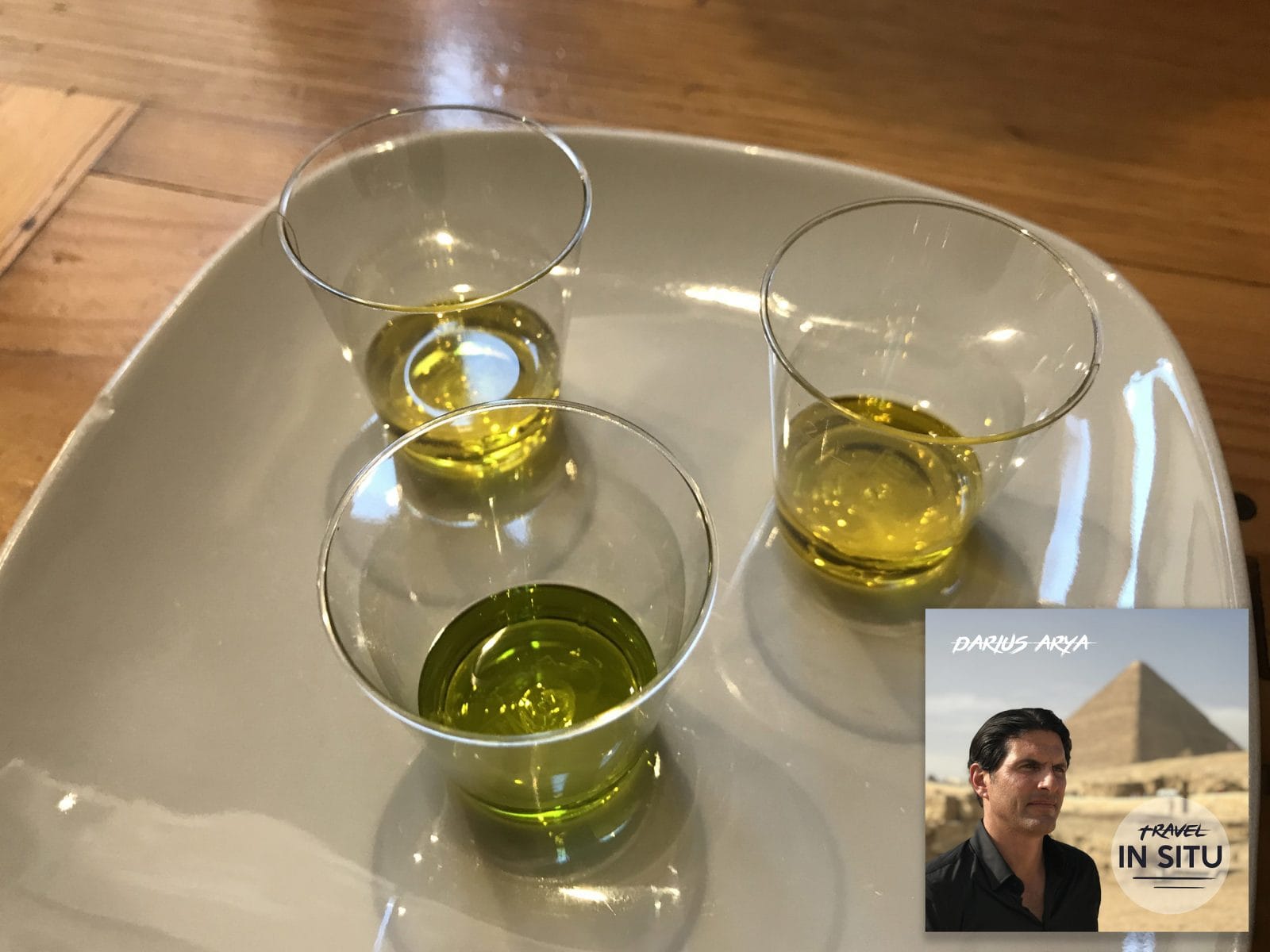 Three small plastic cups filled with olive oil sit on a white plate; a book titled “Travel In Situ” with a man and pyramid is overlaid in the corner.