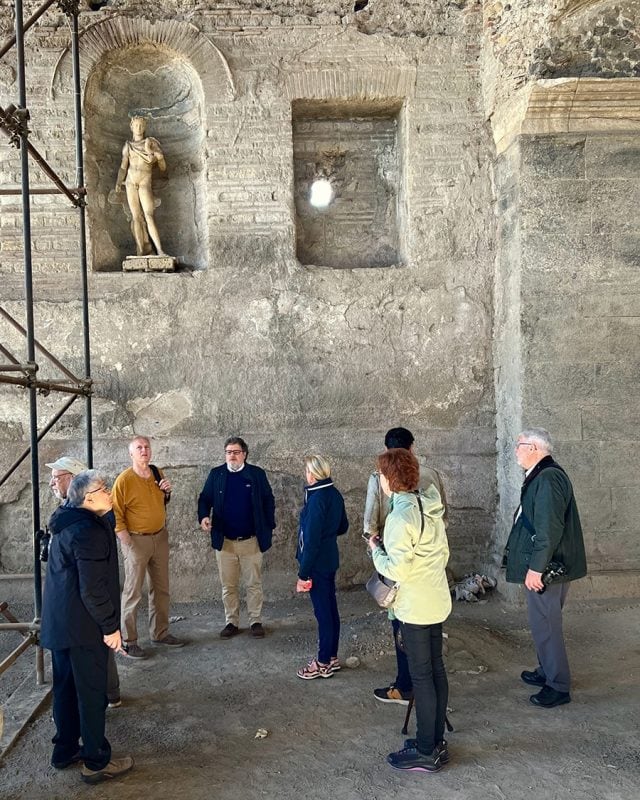 A group of tourists stands in an ancient stone building with a statue of a figure in a wall niche, viewed alongside construction scaffolding.