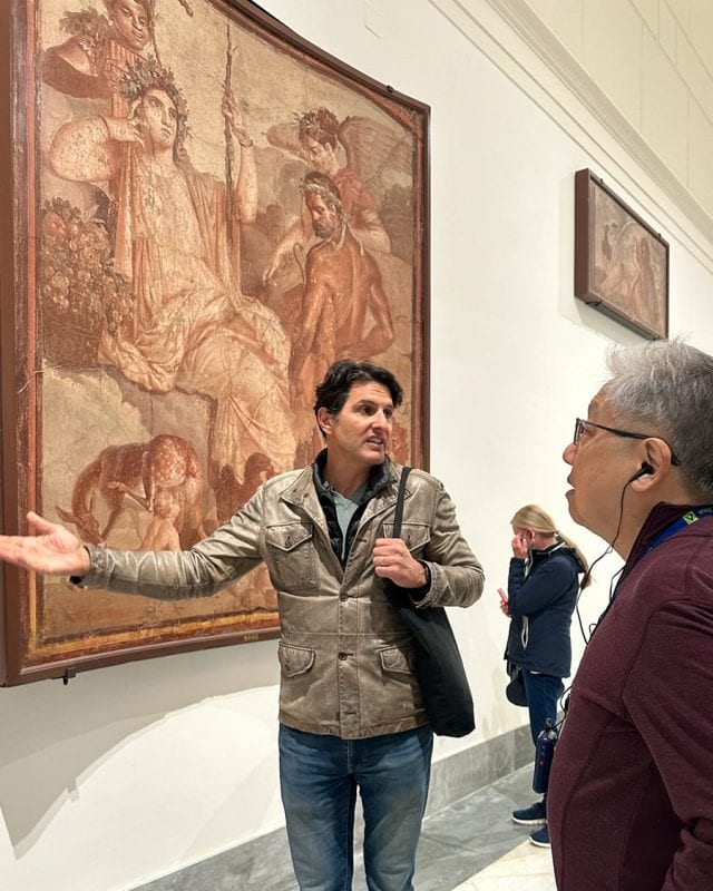 A man in a leather jacket gestures toward a large framed Roman fresco, explaining it to another man in a museum gallery.