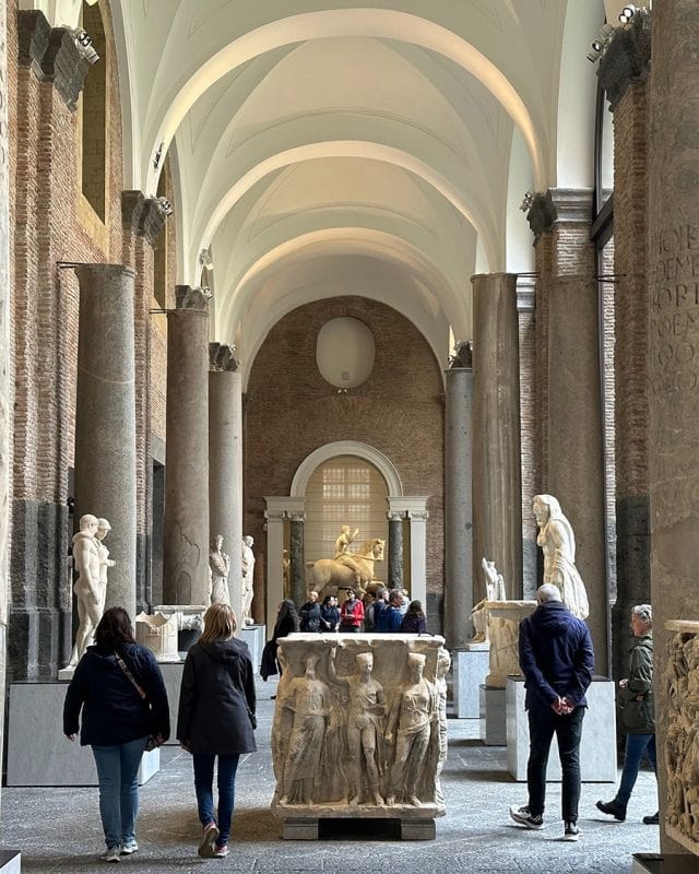 Visitors walk through a vaulted gallery containing multiple marble statues and a relief-carved stone sarcophagus.