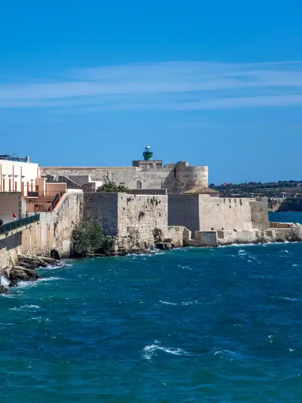 Stone fortress with a lighthouse on top stands by the blue sea under a clear sky. Buildings line the shoreline.