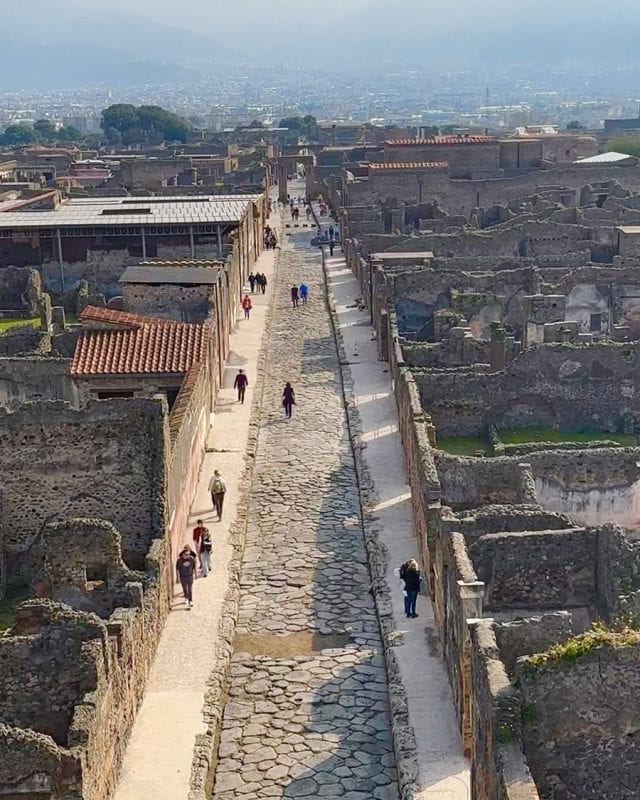 Visitors walk along a stone-paved street running through the ancient ruins of Pompeii, with a sprawling city visible in the background.