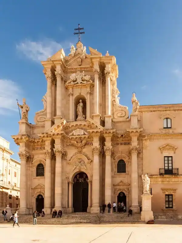 Baroque-style cathedral with ornate facade, statues, and large doors, set against a clear blue sky with people sitting on the steps in front.
