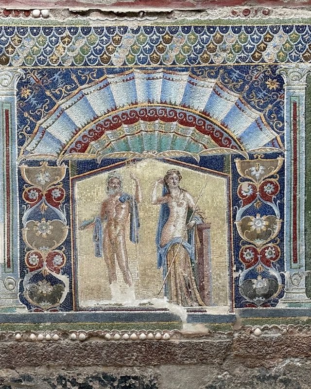Roman wall mosaic depicting a man and a woman standing beneath a decorated arch, framed by ornate floral patterns and a scalloped border.