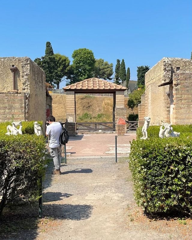 A person takes a photo in an outdoor archaeological site with stone walls, a tiled roof structure, and small statues set among garden hedges.