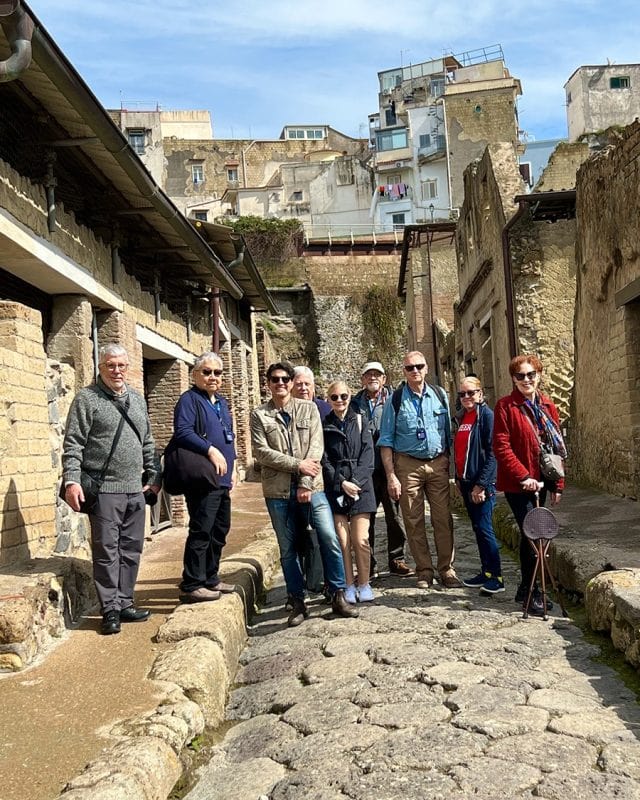 A group of nine tourists walking along a narrow stone-paved street in the ruins of Herculaneum.