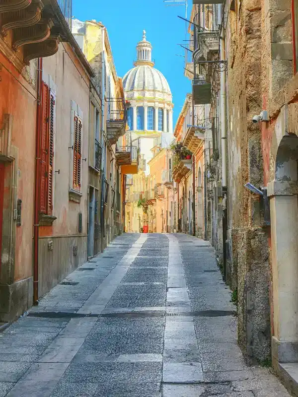 Narrow cobblestone street lined with old buildings, leading to a large domed church under a clear blue sky.