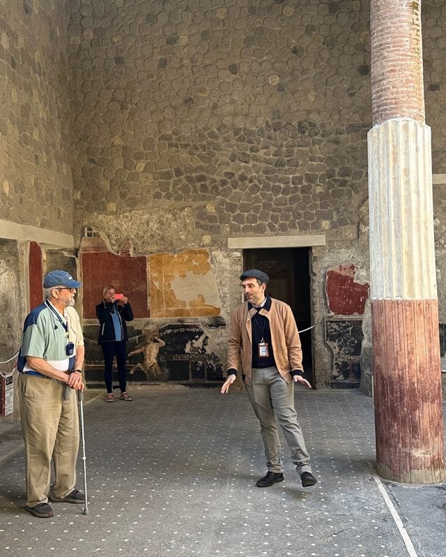 A guide gestures while speaking to a man with a cane and a woman taking a photo inside a ruin with stone walls, painted frescoes, and a large column.