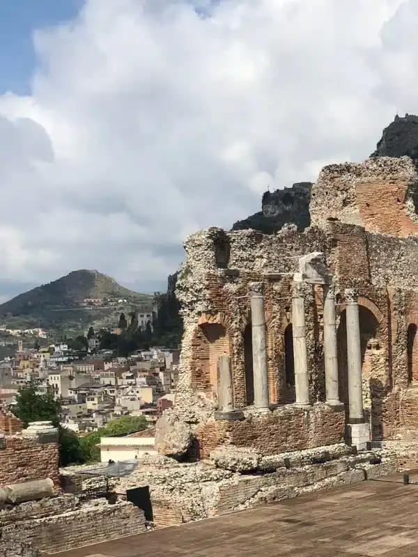 Ancient stone ruins with columns overlook a town and mountains under a partly cloudy sky.