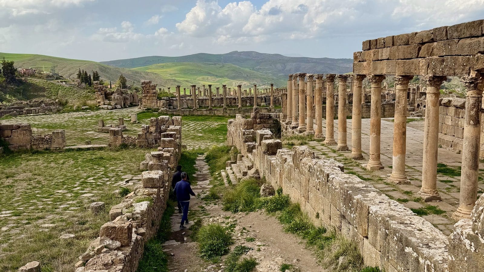 Two people walk through the ruins of the Roman city of Cuicul, featuring rows of standing stone columns and grassy courtyards under a cloudy sky.
