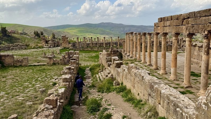 Two people walk through the ruins of the Roman city of Cuicul, featuring rows of standing stone columns and grassy courtyards under a cloudy sky.