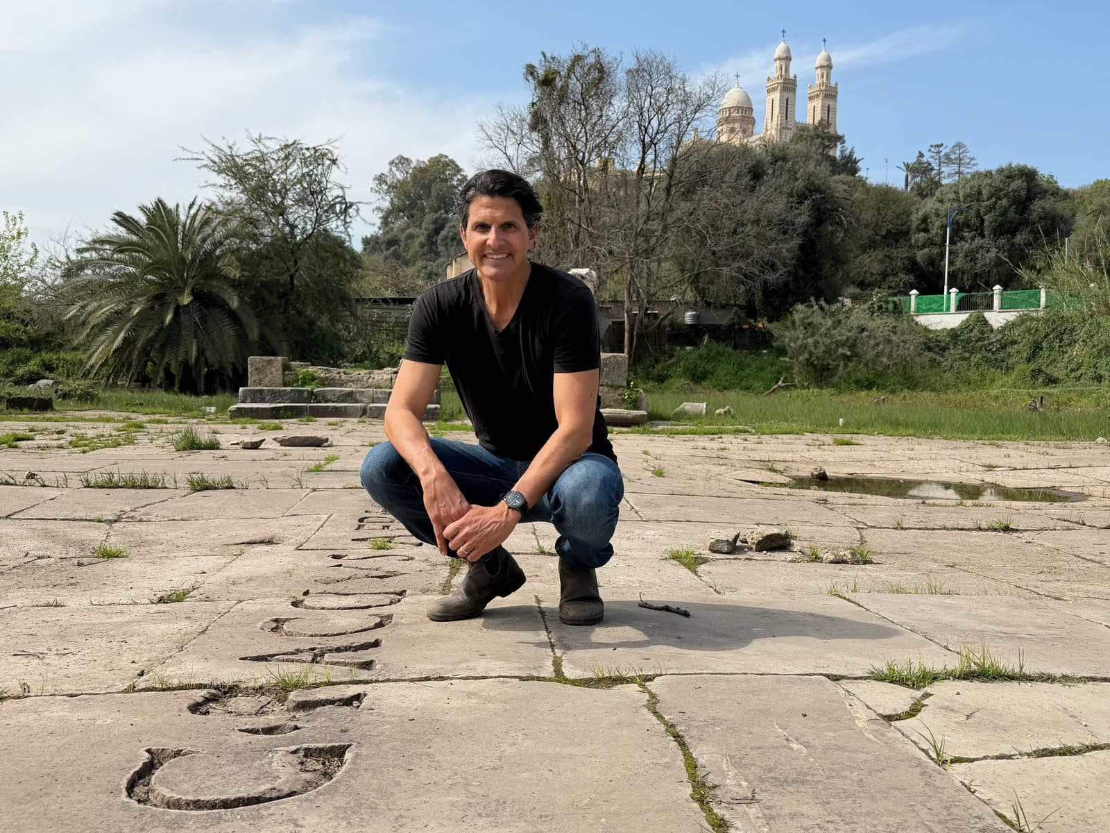 A man in a black t-shirt and jeans crouches on stone ruins with a domed building in the background.