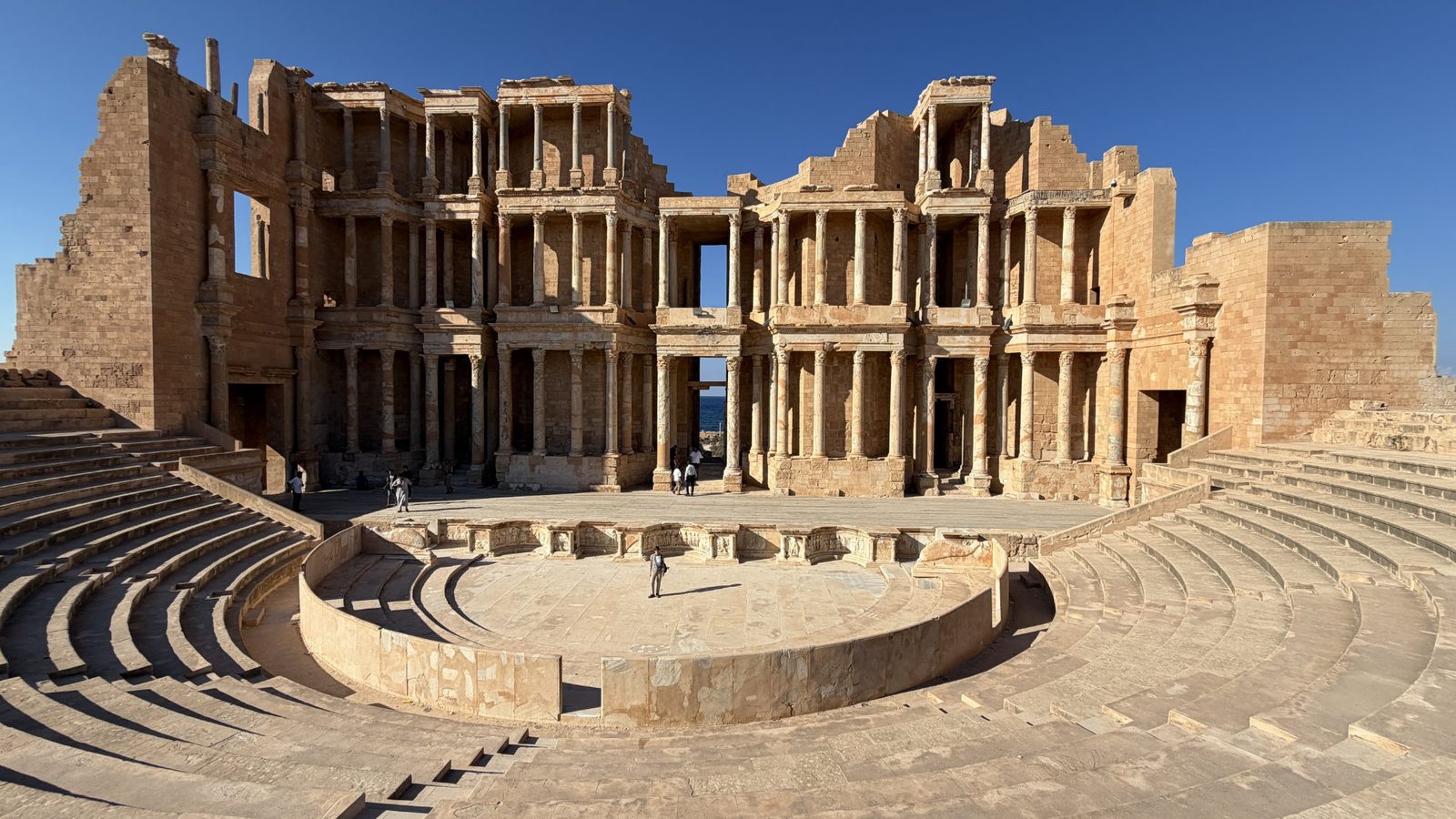 The stone ruins of the Roman theater at Leptis Magna in Libya, featuring a large semicircular stage and tiered seating.