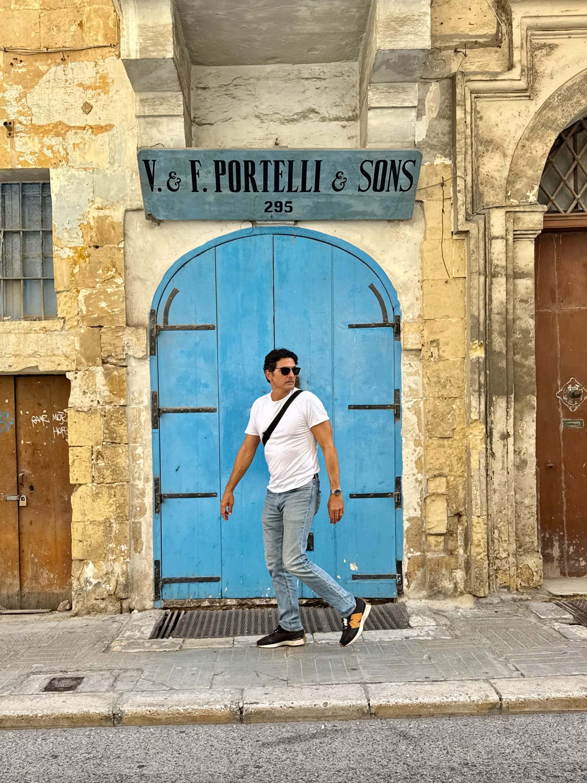 A person in sunglasses and casual clothes walks past a large blue door set in an old stone building with a “V. & F. Portelli & Sons” sign above.