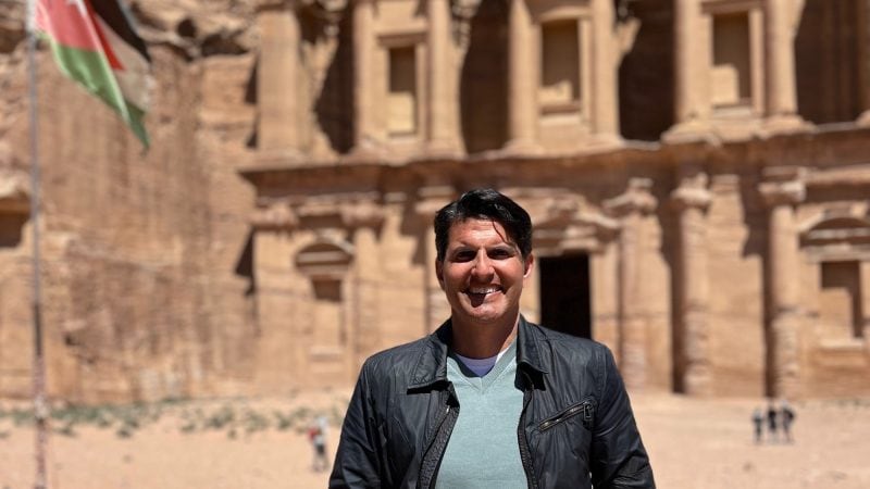 A man smiling in front of the Monastery at Petra, with the Jordanian flag flying to his left.
