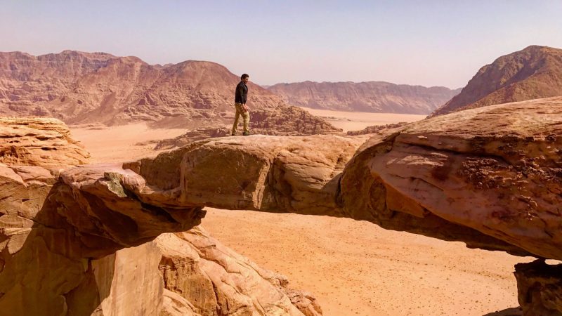 A person stands on a natural sandstone rock bridge overlooking a desert landscape.