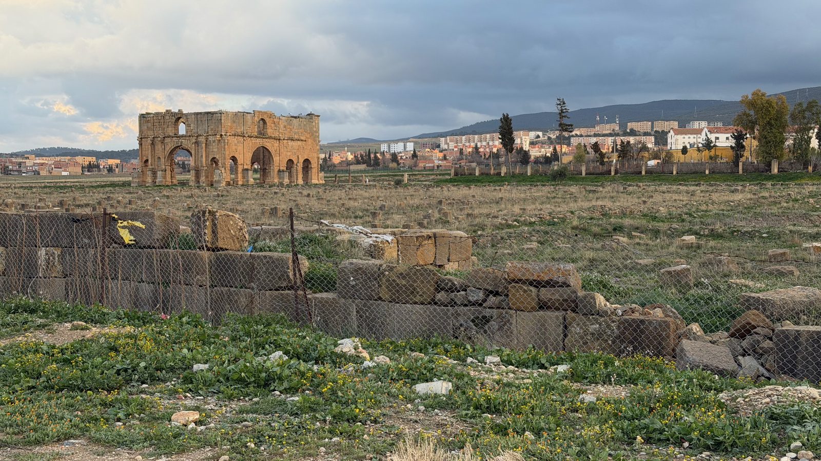 Ancient Roman ruins of an arch standing in a field in front of a modern city and cloudy sky.