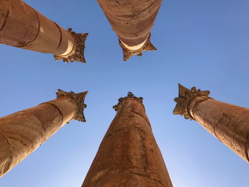 Low-angle view of four ancient stone columns with ornate capitals reaching toward a clear blue sky.