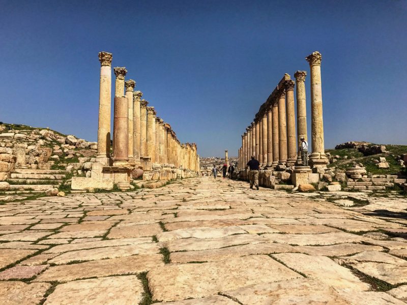 A wide, stone-paved street lined with rows of ancient Roman columns under a clear blue sky, with several people walking between them.
